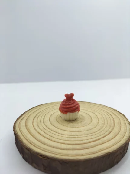 A close-up shot of a miniature cup cake product with pink frosting and a red cherry on top, sitting on a tiny white plate.